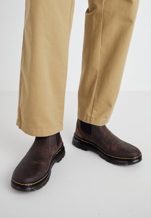 Bruine leren Chelsea-boots met zwarte elastische zijkanten en geelgestikte details, gedragen met beige broek met rechte pijpen.