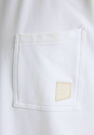 Hvid bomuldsskjorte med en stor lomme med lap, der har et beige stofmærke med et enkelt broderet design og synlige syninger.