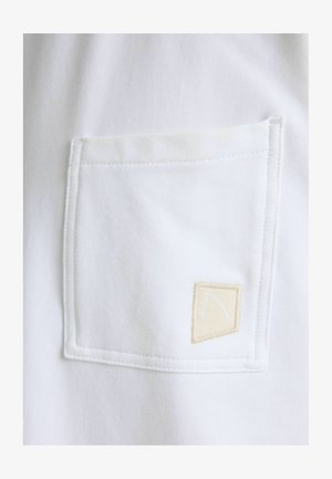 Fehér pamut ing nagy zsebfedővel, bézs szövetcímkével, amely egyszerű hímzett mintát és látható varrást tartalmaz.