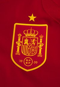 Maglia rossa con un emblema a scudo giallo. Il stemma include una corona, un castello, un leone e del testo su una fascia. I dettagli sono evidenziati in un giallo brillante.