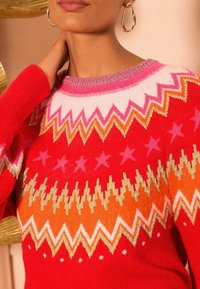 Roter Pullover mit Rundhalsausschnitt, der bunte Zickzackmuster in Pink, Orange und Gold aufweist. Sanfte Textur mit Sternmotiven überall.