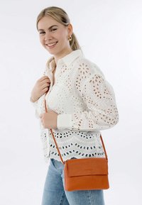 Braune Leder-Umhängetasche mit strukturiertem Finish und Klappenverschluss, getragen mit einem verstellbaren Gurt über einer weißen Lochstickbluse.