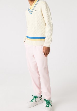 Person trägt cremefarbenen V-Ausschnitt-Pullover mit Zopfmuster und blauen sowie gelben Streifen, blassrosa Hose und weiße Sneaker mit grünen Schnürsenkeln.