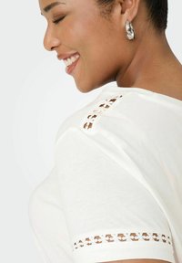 Camiseta blanca de corte relajado, con delicados detalles de encaje en el hombro y mangas cortas. Textura de algodón suave.