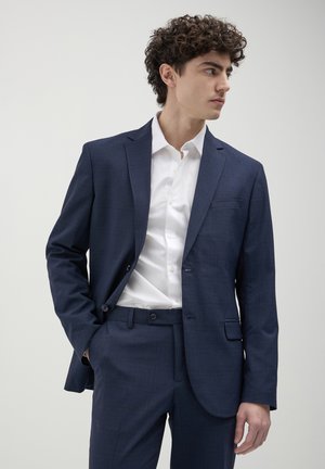 Giovane uomo che indossa un completo blu navy con una camicia bianca, con una mano in tasca, che guarda di lato contro uno sfondo chiaro e uniforme.