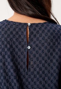 Blusa blu navy ricamata con motivi floreali, apertura a oblò sul retro e due bottoni circolari sul colletto. Dettaglio del tessuto testurizzato visibile.