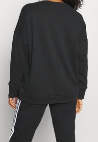 Sudadera negra de corte holgado, con escote ancho y hombros caídos. Tejido suave con puños y dobladillo acanalados. Combinada con pantalones negros que tienen rayas blancas.
