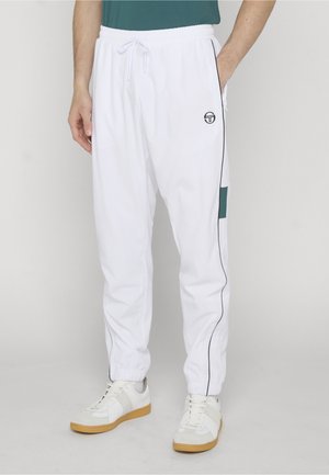 Mężczyzna w białych spodniach dresowych ze sznurkiem w talii, bocznymi lampasami i małym logo, w połączeniu z białymi trampkami na gumowej podeszwie.