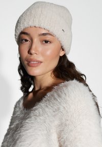 Cream-colored knitted beanie with a ribbed design and a small metallic logo. Soft, textured yarn creates a cozy appearance.