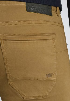Beige Baumwollhose mit einer Gesäßtasche, sichtbaren Nähten, einem Logoetikett über der Tasche und einer glatten, matten Textur.