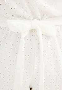 Mono de tela blanca con ojales recortados, que presenta un cinturón atado en la cintura. Textura suave, material liviano, diseño de ajuste relajado.