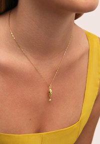 Collar de cadena de oro con un colgante de pez que presenta acentos triangulares en azul y turquesa, colgando sobre piel desnuda por encima de una prenda amarilla.