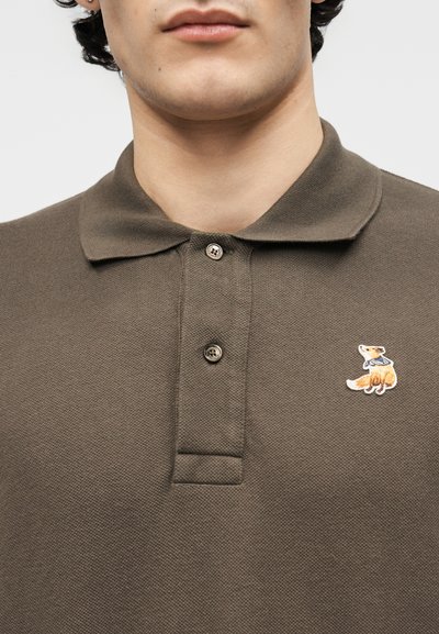 Άνδρας που φοράει ελιάς-πράσινο πόλο μπλουζάκι με δύο κουμπιά κουμπωμένα και μικρή κεντημένη αλεπού με μπλε κασκόλ στο στήθος.