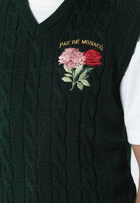 Vihreä neulottu liivi V-kaula-aukkolla, jossa on punoksia ja kirjailtuja kukkia vaaleanpunaisena ja punaisena, sekä kultaiset "PAS DE MONACO" -tekstit.