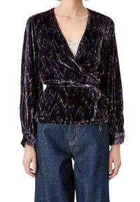 Top avvolgente in velluto di colore viola scuro con motivi multicolori, dotato di maniche lunghe, scollatura a V e un fiocco in vita. Abbinato a jeans.