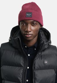 Czarna kamizelka puffer z kapturem, w zestawie z burgundową, prążkowaną czapką beanie z naszywką marki. Tekstura dzianiny i stonowane kolory.