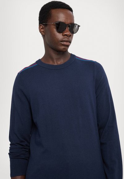 Maglione a maniche lunghe blu navy in morbido tessuto knit con strisce multicolori sulla spalla e scollatura a girocollo. Occhiali da sole rotondi.