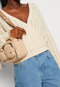 Beige knitted sweater with a V-neck and diamond pattern, paired with a fuzzy beige handbag featuring a front pocket and accent hardware.