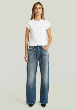 Giovane donna in piedi con una mano in tasca, indossa una semplice t-shirt bianca e jeans blu sbiaditi a gamba larga su uno sfondo neutro.