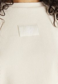 Top de tela crema acanalada que presenta una etiqueta blanca con el logo "SikSilk". Primer plano del escote y la textura con rayas horizontales visibles.