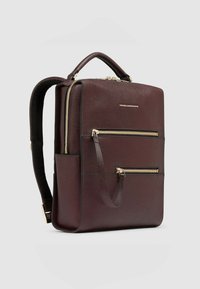 Zaino in pelle color marrone scuro con cerniere dorate, forma strutturata, manico superiore, due tasche frontali con cerniera e spallacci regolabili. Texture liscia.