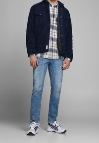 Marine fleecejack met twee borstzakken, laag over een geruit shirt, gecombineerd met lichtblauwe spijkerbroek en witte sneakers met zwarte accenten.