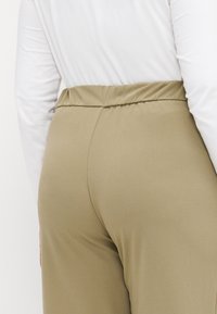 Elastisk liv, bukser i khaki-farge med en glatt tekstur, og en samlet midje. Materialet er mykt og former seg etter kroppen.