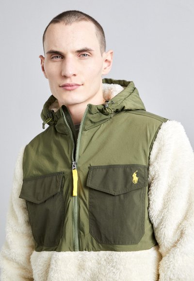 Žalia zip-up striukė su smėlio spalvos kailio tipo rankovėmis. Yra geltonas užtrauktuko traukiklis, dvi priekinės kišenės ir mažas logotipas ant krūtinės.