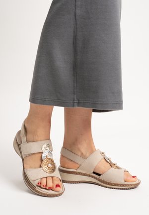 Feet wearing beige wedge sandals with pearl and metallic circular decorations, paired with a knee-length gray skirt.