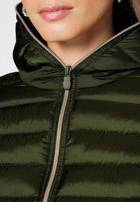 Grøn pufferjakke med lynlåsåbning, der har vandret quiltede sektioner og hætte. Glat tekstur med en kontrasterende lynlås.