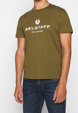 Homme portant un t-shirt à manches courtes col rond couleur vert olive avec le texte et le logo "BELSTAFF ENGLAND 1924", associé à un jean bleu foncé.