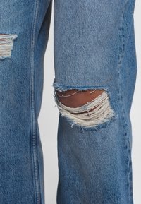 Jeans in denim blu distressed con ampie strappi sfilacciati sulle ginocchia. Presentano una vestibilità ampia e un tessuto testurizzato. Cuciture visibili e bordi grezzi.