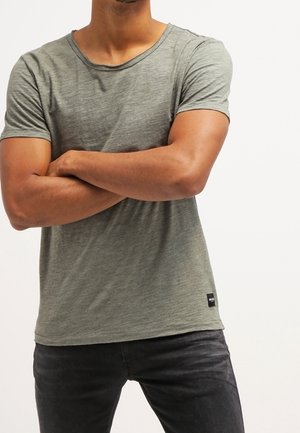 Mężczyzna w dopasowanej szarej koszulce z krótkim rękawem, ze skrzyżowanymi ramionami, w czarnych dżinsach, stojący na jednolitym tle.