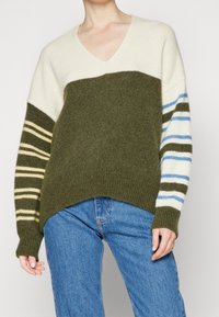 Sweter w serek w oliwkowym zielonym i kremowym kolorze, z paskowanymi rękawami w ciemnozielonym, kremowym i jasnoniebieskim odcieniu. Miękka faktura i luźny krój.