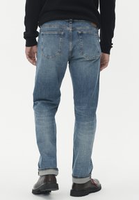 Asmuo vilkintis šviesiai mėlynas, sukurtas iki kulkšnies džinsus, juodą ilgomis rankovėmis marškinėlį ir tamsiai rudas batus su raudonomis raištelėmis, stovintis atsisukęs atgal ant baltos fono.