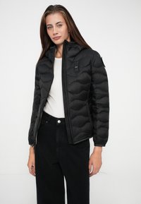 Black padded jacket with a high collar, zip closure, and textured shell. Pairing with a light shirt and black pants. Features a logo patch.