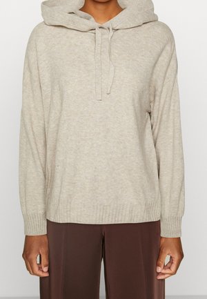 Persoon draagt een licht beige gebreide hoodie met trekkoorden en lange mouwen, gecombineerd met donkerbruine broek, staand tegen een effen achtergrond.