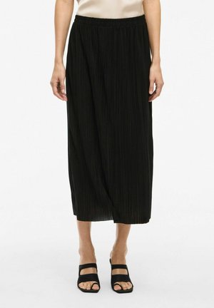 Jupe plissée noire avec une taille élastique, longueur mi-mollet et texture lisse. Associée à des sandales noires à bout ouvert.