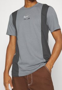 Šedé tričko Nike s tmavě šedými bočními panely, kulatým výstřihem a vyšitým logem "AIR" na hrudi v barvě stříbra. Krátké rukávy, směs bavlny.