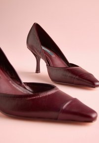 Scarpe con tacco alto in pelle borgogna con punta affilata, caratterizzate da una finitura testurizzata e un taglio curvo a metà piede.