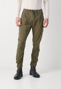 Olivengroene cargobroek met een elastische tailleband, aantrekkoord, zijzakken en aansluitende boorden. Draagt met zwarte enkellaarsjes.