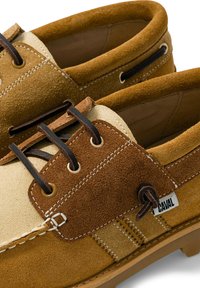 Gros plan sur des chaussures bateau en daim marron et beige avec des lacets noirs et des coutures blanches, avec une petite étiquette blanche indiquant la marque Caval.