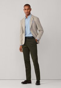 Mann trägt beigen Blazer, hellblaues Hemd, dunkelgrüne Hose, schwarzen Gürtel und dunkelbraune Tassel-Loafers, steht vor einfarbigem Hintergrund.