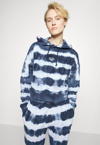 Sweater com efeito tie-dye azul e branco, apresentando um bolso frontal, capuz ajustável com cordão e punhos canelados. Feito de um tecido macio e texturizado.