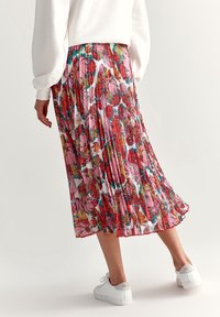 Gonna lunga plissettata in un vivace motivo floreale con tonalità di rosa, rosso e verde. Tessuto leggero, design fluido, indossata con sneakers bianche.