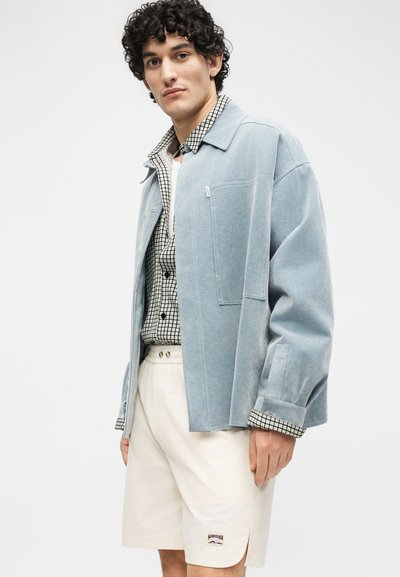 Jeune homme aux cheveux bouclés portant une veste oversize bleu clair, une chemise à carreaux, un t-shirt blanc et un short crème avec un logo.