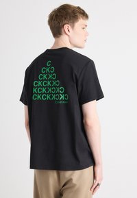 Svart bomull T-skjorte med grønn grafisk design på baksiden, som viser gjentatte bokstaver og merkevarenavnet "Calvin Klein" nederst.