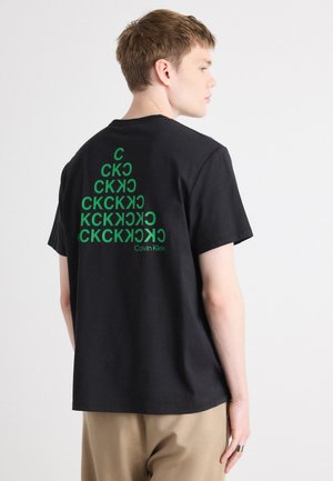 Crna pamučna majica s zelenim grafičkim dizajnom na leđima, koja prikazuje ponavljajuća slova i naziv brenda "Calvin Klein" na dnu.