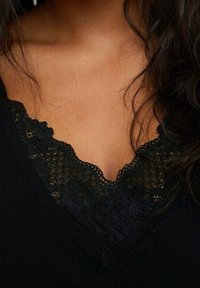 Top nero con scollatura a V profonda, rifinito con delicata pizzo nero, che evidenzia un motivo in pizzo testurizzato su uno sfondo di tessuto liscio.