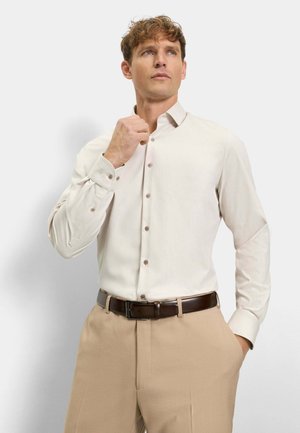 Ein Mann, der den Kragen seines beigen Hemdes mit Knöpfen einstellt, trägt hellbraune Hosen und einen dunkelbraunen Gürtel, steht vor einem einfarbigen hellen Hintergrund.